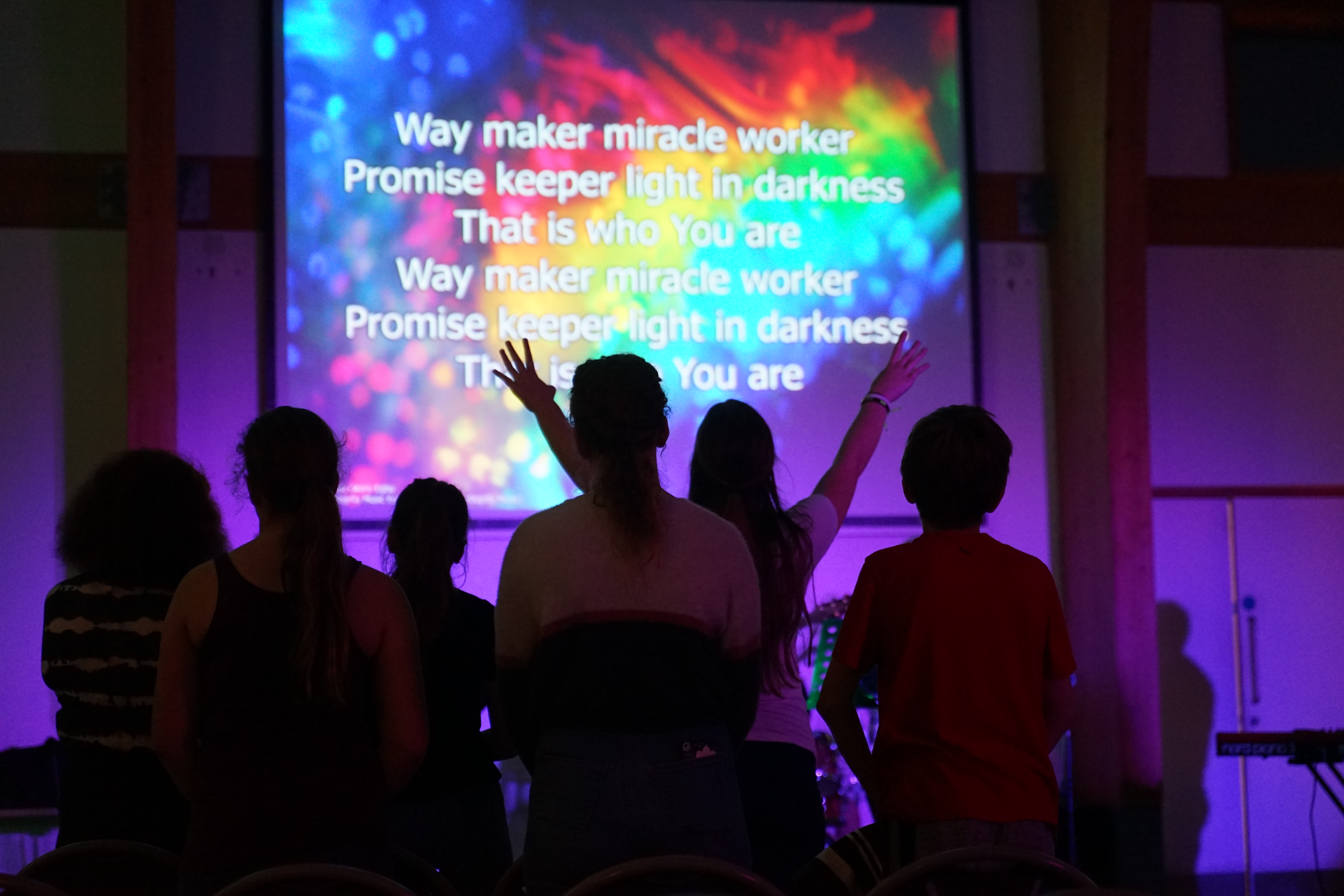 Young people worshipping in church, they have their backs to the camera and have a screen in front of them displaying song words on a coloured background